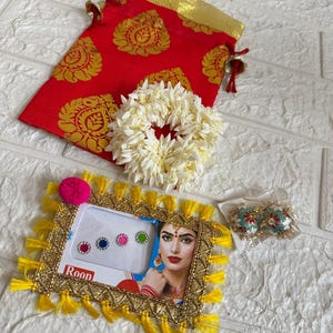 May include: An assortment of Indian-inspired accessories. Includes a red pouch with gold floral patterns, a white flower garland, a framed picture with colorful bindi stickers, and a pair of floral earrings.