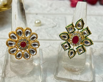 May include: Two gold-toned rings with intricate floral and geometric designs. Each ring features a central red gemstone surrounded by clear and green accents. The rings are displayed on clear stands, showcasing their detailed craftsmanship.