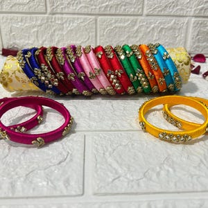 May include: A set of colorful silk thread bangles with decorative embellishments. The bangles are arranged in a stack and are displayed on a white surface.