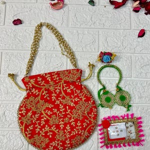 May include: A red embroidered pouch with a gold chain handle, a green and gold bangle, a flower hair clip, and a pink frame with a small mirror and a picture of a woman.