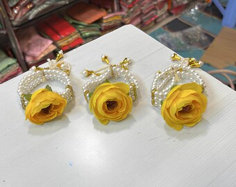 May include: Three floral wrist corsages. Each corsage features a yellow rose and a white pearl bracelet. The corsages are adorned with gold accents. The items are displayed on a white surface.