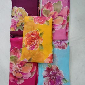 Op de afbeelding: Een verzameling kleurrijke stoffen met bloemenprint in tinten paars, roze, geel en blauw. Elke stof heeft een groot, gedetailleerd bloemmotief met zilveren accenten. De stoffen zijn netjes opgevouwen en gestapeld.