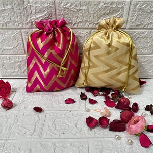 Può includere: Due sacchetti decorativi con coulisse e motivo a chevron. Un sacchetto è rosa con dettagli dorati e l'altro è dorato con dettagli dorati.
