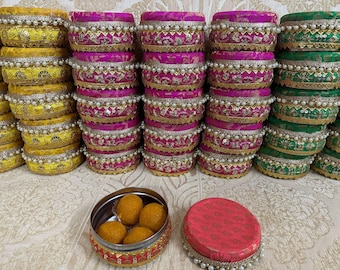 Op de afbeelding: Een assortiment decoratieve, ronde containers in geel, fuchsia en groen, gestapeld in rijen. Elke container is versierd met gouden randen, witte parels en ingewikkelde patronen. Een kleine, open metalen container bevat verschillende oranje snoepjes, met een bijpassende deksel.