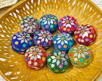 May include: A collection of ten small, round, decorative boxes with mirrored centers and colorful beaded embellishments. The boxes are arranged in a circular pattern in a woven basket.