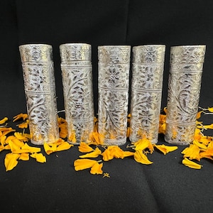 May include: Five silver cylindrical containers with intricate floral patterns. The containers are arranged in a row, with bright yellow flower petals scattered around them. The background is black.