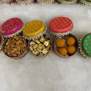 Puede incluir: Una colección de recipientes redondos decorativos con tapas. Los recipientes están llenos de almendras, bombones y dulces redondos de color naranja. Las tapas están cubiertas de tela roja, rosa, amarilla y verde, y adornadas con perlas.