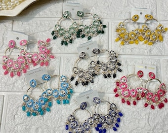 May include: A collection of hoop earrings in various colors, including pink, green, yellow, black, blue, and red. Each pair features a circular design with decorative stones and dangling beads. The earrings are displayed on a white surface.