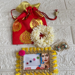 May include: An assortment of Indian-inspired accessories. A red and gold drawstring bag, a white flower garland, a framed picture with a woman's image, and a pair of earrings. The frame has yellow tassels and the word "Roop".