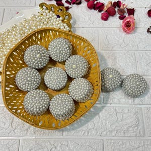 May include: A wicker basket filled with small, round, white pearl-covered boxes. The boxes are arranged in a circular pattern and are surrounded by white pearl beads.