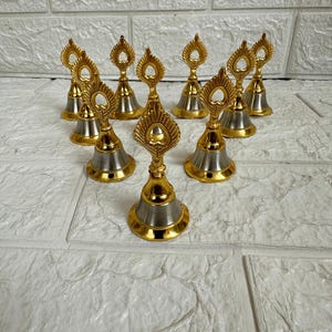 May include: A set of ten small, gold and silver bells with a peacock feather design. The bells are arranged in a circle on a white tiled surface.