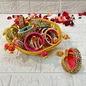 May include: A collection of colorful silk thread bangles with gold accents and embellishments. The bangles are arranged in a woven basket with a few loose bangles on the white surface.
