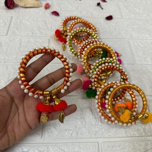 May include: A collection of colorful bangles. The bangles are adorned with white pearls, gold accents, and red, orange, green, and yellow tassels. One bangle is held in a hand, showcasing the red and gold design with leaf charms.