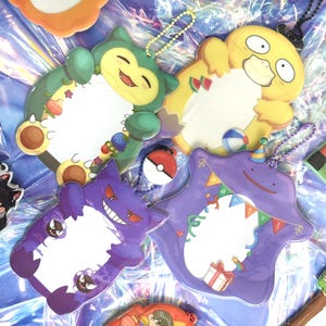 May include: A collection of four acrylic photo frames shaped like cartoon characters including a green Snorlax, a yellow Psyduck, a purple Gengar, and a purple Ditto wearing a party hat, each featuring a central white space and a ball chain.