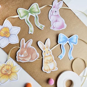 May include: An Easter garland with watercolour illustrations of bunnies, bows, and daffodils. The garland includes a pink bunny, a brown bunny, and a yellow bunny. The bows are green and blue. The daffodils are white and yellow.