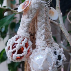 May include: A macrame keyring featuring three spheres encased in off-white braided cord netting. The spheres include one translucent orange glass orb, one opaque white orb, and one dark textured orb. Small white seashells are attached to the metal ring.