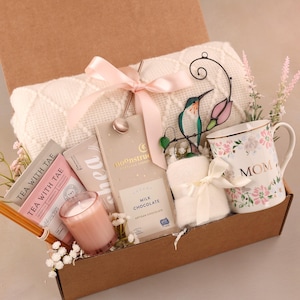 May include: A gift box filled with a white blanket, a floral "MOM" mug, tea packets, a candle, chocolate, and a decorative hummingbird. The box is tied with a pink ribbon and includes decorative flowers.