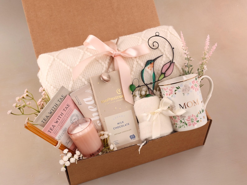 May include: A gift box filled with a white blanket, a floral "MOM" mug, tea packets, a candle, chocolate, and a decorative hummingbird. The box is tied with a pink ribbon and includes decorative flowers.