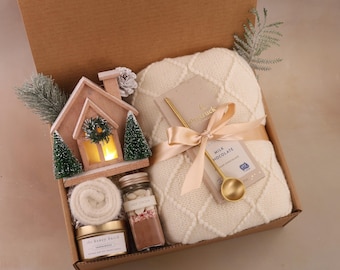 May include: A gift box filled with cozy items. Includes a cream-colored knitted blanket, a miniature wooden house with a lit window, a candle, hot cocoa mix, and a gold spoon. The box is tied with a cream ribbon.