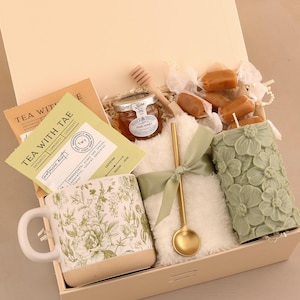May include: A gift box filled with tea, honey, a floral mug, a candle, and caramels. The mug and candle are green. The box contains a gold spoon and a small jar of honey. The tea packaging reads "TEA WITH TAE".