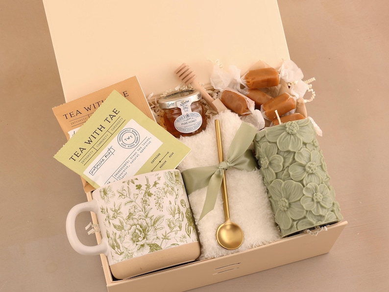 May include: A gift box filled with tea, honey, a floral mug, a candle, and caramels. The mug and candle are green. The box contains a gold spoon and a small jar of honey. The tea packaging reads "TEA WITH TAE".