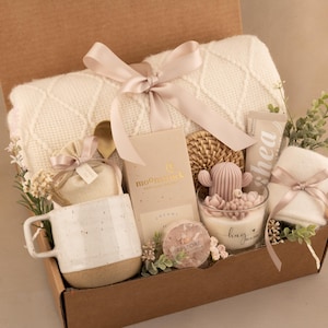 May include: A gift box filled with a cream-colored blanket, a white mug with a speckled base, a cactus-shaped candle, and other self-care items. The box is tied with a pink ribbon and decorated with greenery.