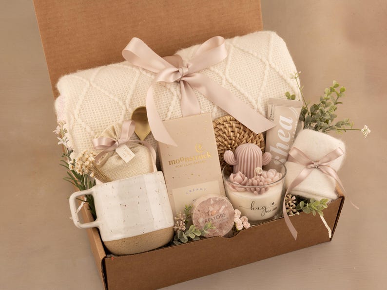 May include: A gift box filled with a cream-colored blanket, a white mug with a speckled base, a cactus-shaped candle, and other self-care items. The box is tied with a pink ribbon and decorated with greenery.