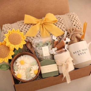 May include: A gift box filled with a white mug that says "BEST MOM EVER", a knitted blanket, crocheted sunflowers, a wooden bowl with daisy candles, soap, and wrapped candies. The box is tied with a yellow bow.