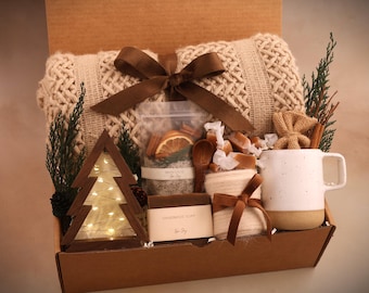 Puede incluir: Una caja de regalo llena de artículos acogedores. Incluye una manta de punto color crema, un árbol de Navidad de madera con luces, una taza blanca, sales de baño, jabón artesanal, caramelos envueltos y un lazo decorativo.