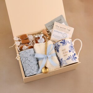 May include: A gift box filled with a floral-patterned candle, a mug with "best MOM ever" text, tea packets, honey, caramels, a small towel, and a golden spoon. The mug has blue floral designs. The box is beige.