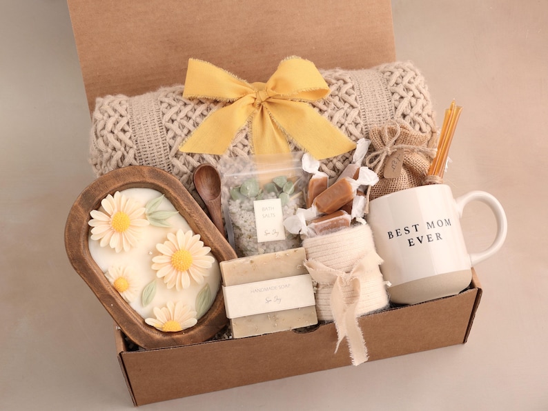 May include: A gift box filled with self-care items. Includes a white mug with "Best Mom Ever" text, a wooden bowl with daisy-shaped soap, a beige blanket, and other treats. The box is tied with a yellow ribbon.