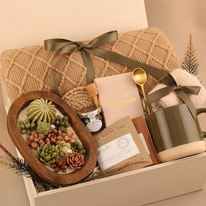 May include: A gift box filled with a beige knit blanket, a wooden bowl with succulent-shaped soaps, a mug, tea, honey, and a gold spoon. The box is tied with a green ribbon.