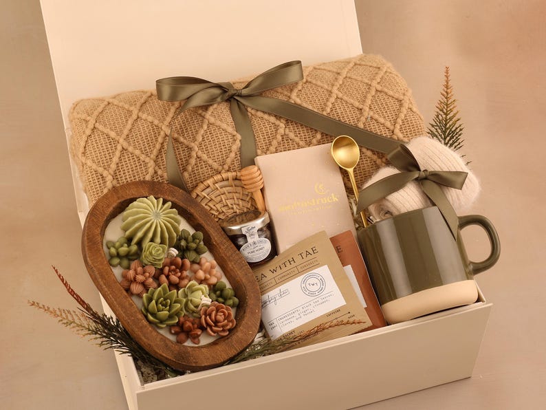 May include: A gift box filled with a beige knit blanket, a wooden bowl with succulent-shaped soaps, a mug, tea, honey, and a gold spoon. The box is tied with a green ribbon.