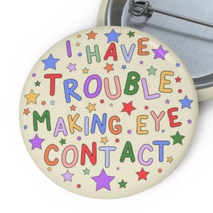 May include: A round button with a white background and colourful text that reads "I HAVE TROUBLE MAKING EYE CONTACT" surrounded by colourful stars.