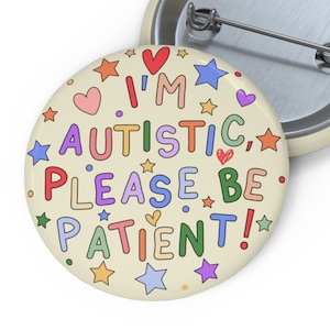 May include: A round button with a cream background and colorful text that reads "I'm autistic, please be patient!" surrounded by stars and hearts.