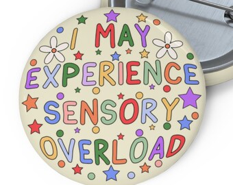 Könnte beinhalten: Ein runder Knopf mit cremefarbenem Hintergrund und buntem Text, der "I MAY EXPERIENCE SENSORY OVERLOAD" lautet. Der Text ist von bunten Sternen und Blumen umgeben.