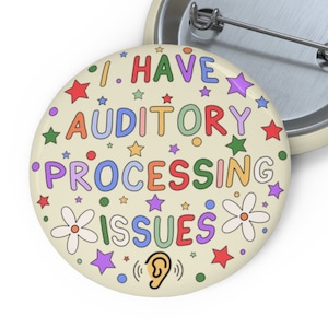 Può includere: Un bottone rotondo con sfondo crema e testo colorato che recita "Ho problemi di elaborazione uditiva". Il testo è circondato da stelle e fiori. C'è una piccola icona di orecchio nella parte inferiore del pulsante.