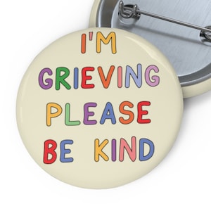 May include: A round button with a cream background and the text "I'M GRIEVING PLEASE BE KIND" in rainbow colours.