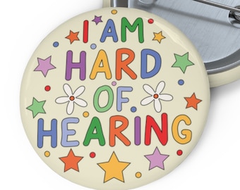 Puede incluir: Un botón redondo con fondo blanco y texto colorido que dice "I AM HARD OF HEARING". El texto está rodeado de estrellas y flores de colores.
