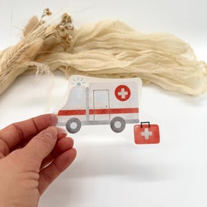 Könnte beinhalten: Ein transparenter Aufkleber mit einem weißen Krankenwagen mit roten Streifen und einem roten Kreuz an der Seite. Der Krankenwagen hat ein blaues Licht oben. Neben dem Krankenwagen befindet sich eine rote Erste-Hilfe-Box mit einem weißen Kreuz.