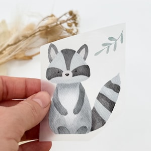 Könnte beinhalten: Eine Aquarell-Illustration eines Waschbären mit einem grau-schwarz gestreiften Schwanz. Der Waschbär sitzt auf einem weißen Hintergrund mit einem kleinen grünen Blattzweig in der oberen rechten Ecke.