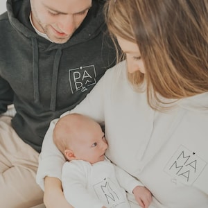 Könnte beinhalten: Eine dreiköpfige Familie trägt passende Hoodies mit den Worten "Papa", "Mama" und "Mini" darauf. Das Baby wird von den Eltern gehalten.