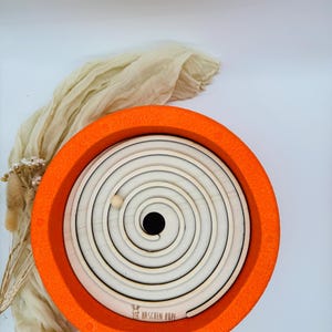 Könnte beinhalten: Ein Holz-Spiral-Labyrinth-Spielzeug mit einem orangefarbenen Rand und einer kleinen Holzkugel. Das Labyrinth hat ein weißes und helles Holz-Spiral-Design. Das Spielzeug hat den Text "HASCHEN HOPF" unten.
