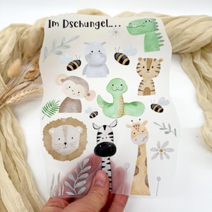 Könnte beinhalten: Ein transparentes, rechteckiges Blatt mit Aquarellillustrationen von Dschungeltieren, darunter ein grünes Krokodil, ein Tiger, ein Zebra, eine Giraffe und ein Löwe. Der Text "Im Dschungel..." ist oben aufgedruckt. Das Blatt wird von einer Hand gehalten.