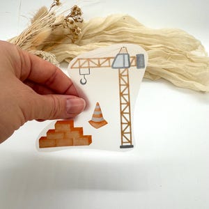 Könnte beinhalten: Ein transparenter Aufkleber mit einem Baustellenthema. Das Design umfasst einen braunen Kran, einen orangefarbenen Verkehrskegel und einen Stapel Ziegelsteine. Der Aufkleber wird von einer Hand vor einem neutralen Hintergrund gehalten.