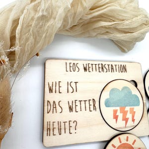 May include: Wooden weather station with the text "LEOS WETTERSTATION" and "WIE IST DAS WETTER HEUTE?" The station includes interchangeable weather symbols, such as a cloud with lightning and a sun with clouds. The station is made of light-colored wood.