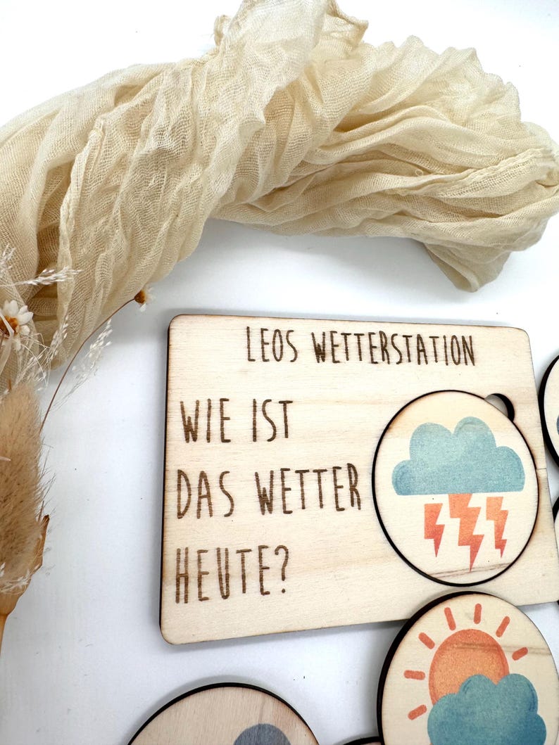 May include: Wooden weather station with the text "LEOS WETTERSTATION" and "WIE IST DAS WETTER HEUTE?" The station includes interchangeable weather symbols, such as a cloud with lightning and a sun with clouds. The station is made of light-colored wood.