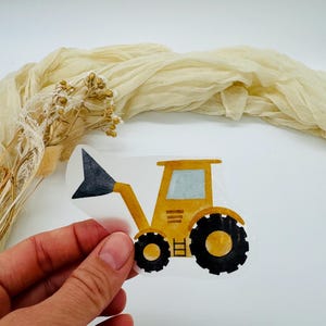 May include: A yellow and black tractor sticker with a raised front loader and large black tires. The sticker is held by a hand against a white background with a beige fabric and dried flowers.