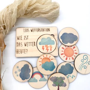 May include: Wooden weather station toy with the text "LEOS WETTERSTATION" and "WIE IST DAS WETTER HEUTE?" The set includes a wooden board with a weather symbol and several round wooden tokens with weather illustrations such as clouds, sun, and rain.