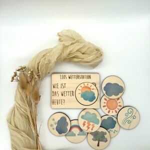 May include: Wooden weather station toy with the text "LEOS WETTERSTATION" and "WIE IST DAS WETTER HEUTE?" includes weather symbols like sun, clouds, rain, and snow. The set has a light wood finish and colorful illustrations.